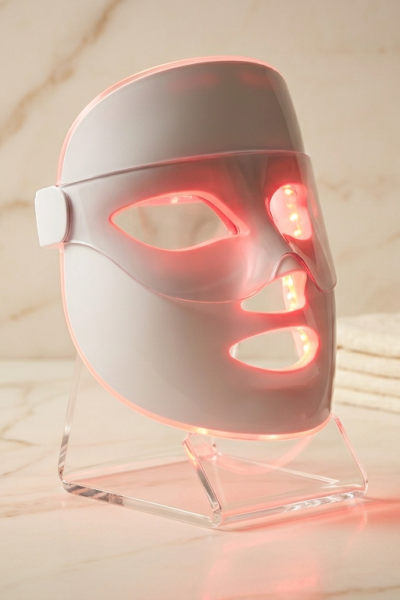 Shark CryoGlow LED face mask glowing red, resting on acrylic stand on marble surface with towel and amber skincare bottle