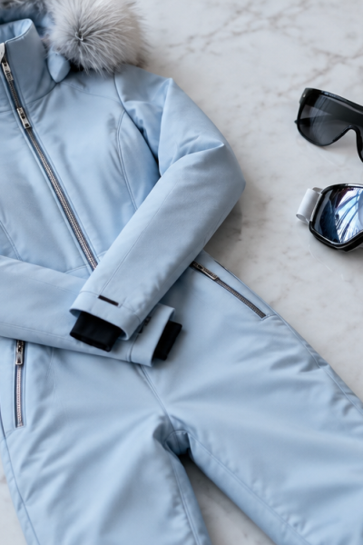 Icy-blue one-piece ski suit, oversized black shield sunglasses, ski goggles, and gold necklace styled on white marble