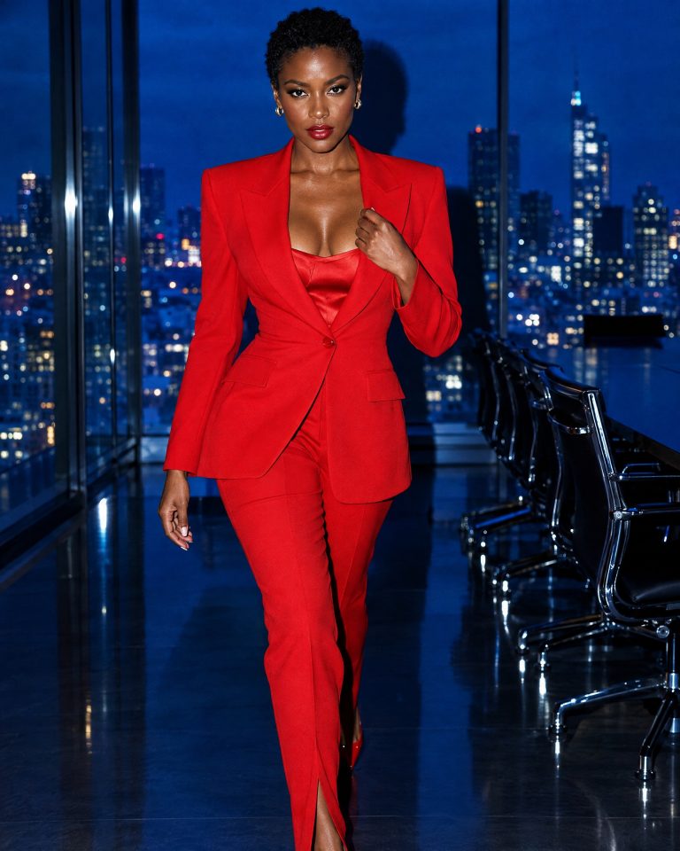 A woman with a short natural crop in a scarlet suit, walking through a dark boardroom with a blue-hour city skyline behind her.
