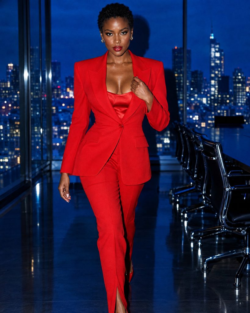 A woman with a short natural crop in a scarlet suit, walking through a dark boardroom with a blue-hour city skyline behind her.