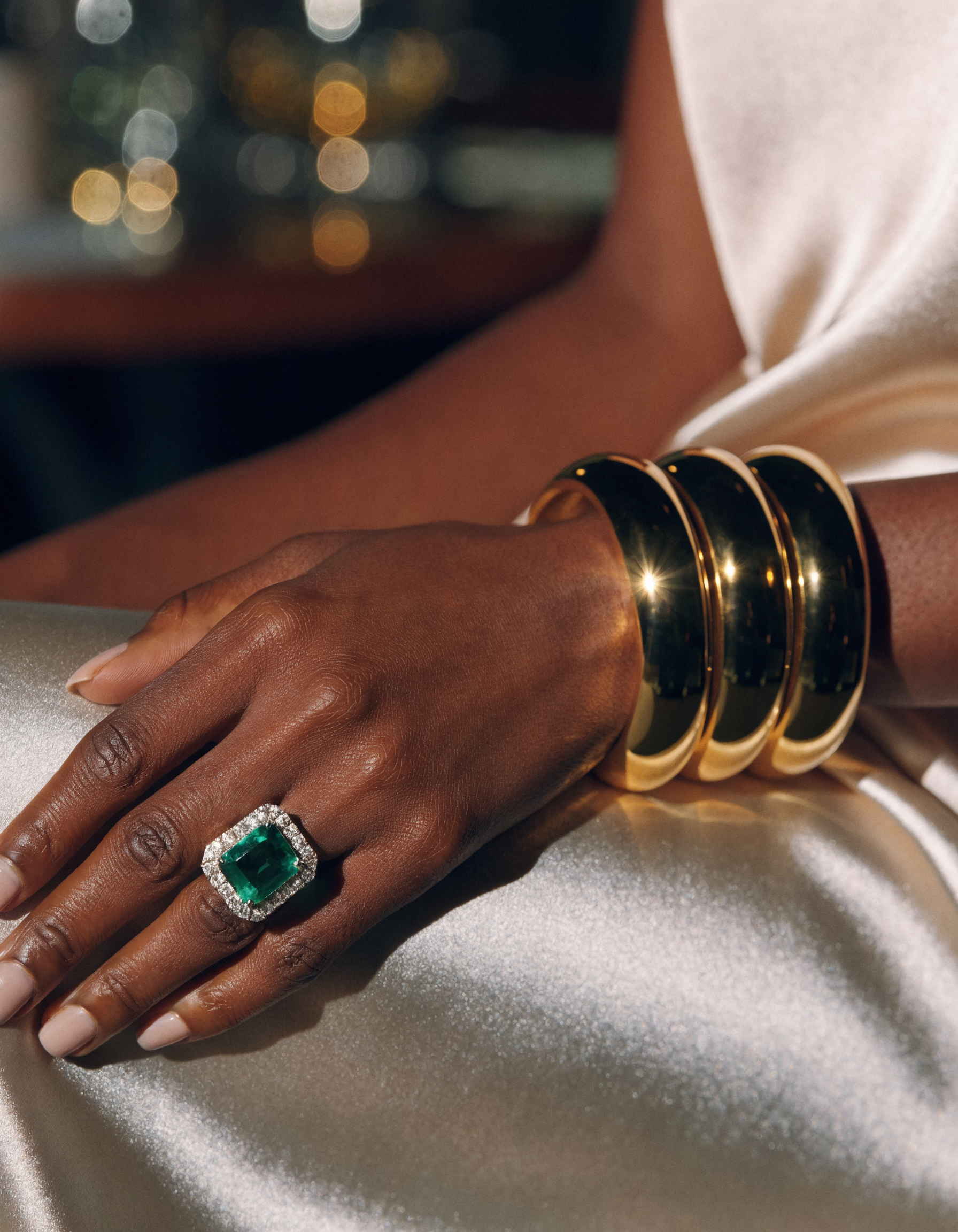 Wrist with stacked gold bangles and an emerald cocktail ring resting on a champagne silk dress